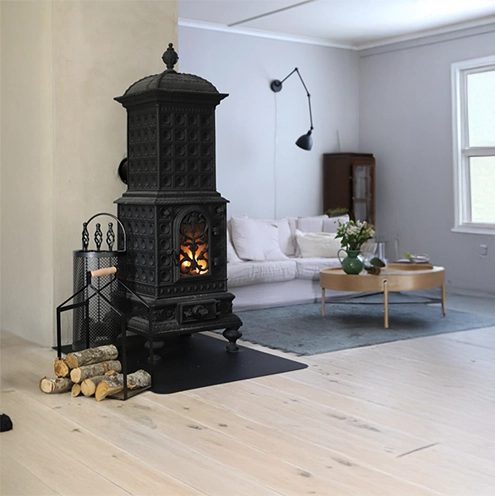 Sortiment 22 En svart gjutjärnskamin med en brasa i står bredvid en stapel ved i ett ljust, modernt vardagsrum med ljust trägolv, en vit soffa, ett runt soffbord och ett stort fönster.