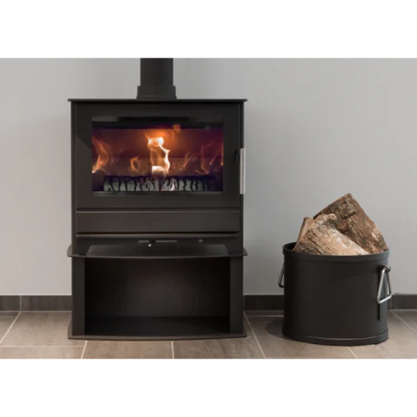 Heta Inspire 55H, en modern svart braskamin med synlig eld inuti, står på ett klinkergolv bredvid en svart metallhink med ved mot en ljusgrå vägg.