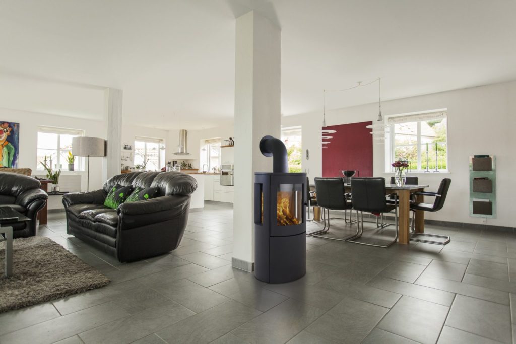 Modernt vardagsrum och matplats i öppen planlösning med svarta skinnsoffor, fristående braskamin, matbord med svarta stolar, stora fönster och klinkergolv.