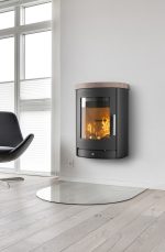 En modern braskamin med glaslucka är monterad på en vit vägg i ett ljust rum med ljust trägolv och stora fönster. En svart stol står i närheten och en glasplatta ligger på golvet under kaminen.