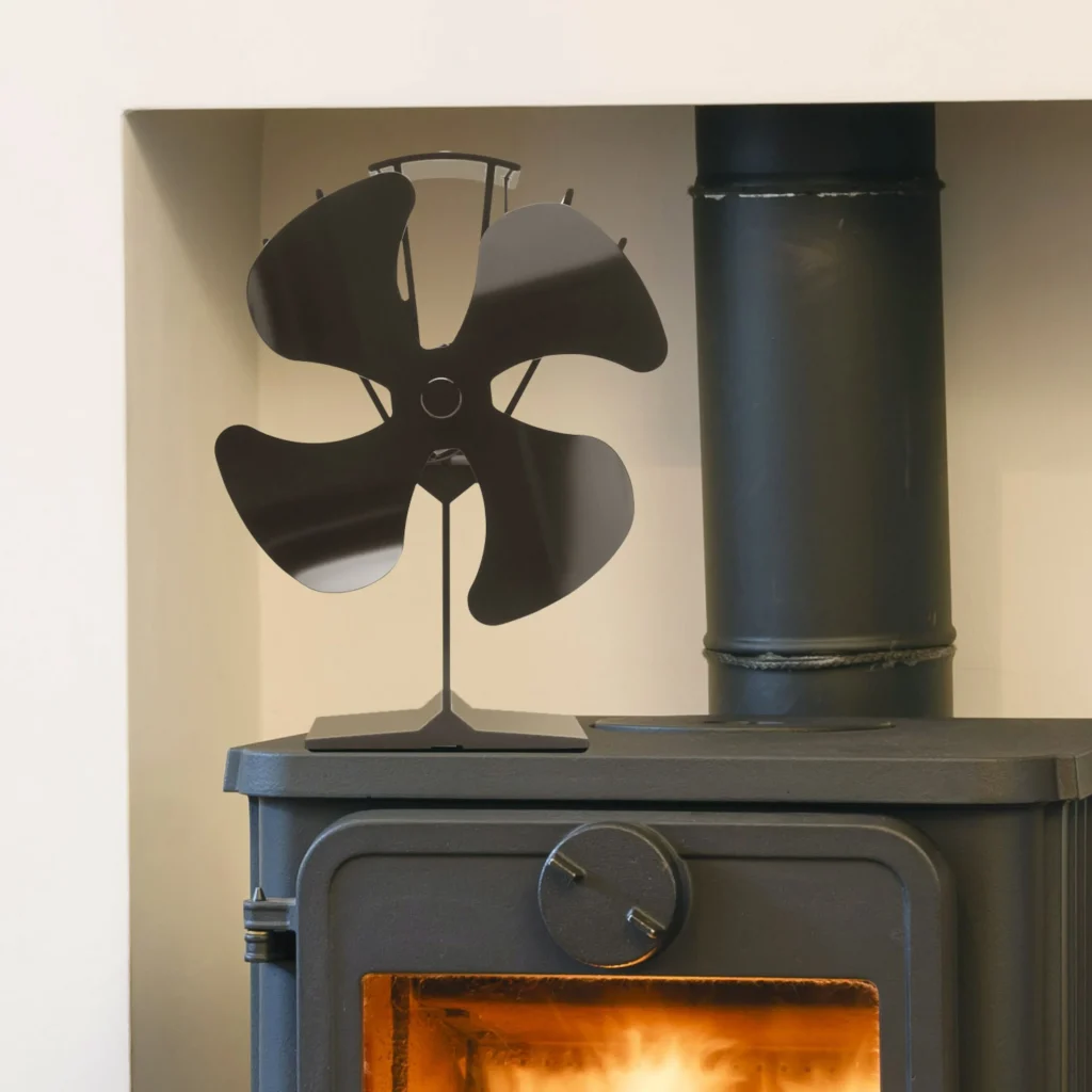 En svart kaminfläkt står ovanpå en vedeldad kamin med en synlig eld som glöder inuti. I bakgrunden syns en vit vägg och ett svart spisrör som sträcker sig uppåt.