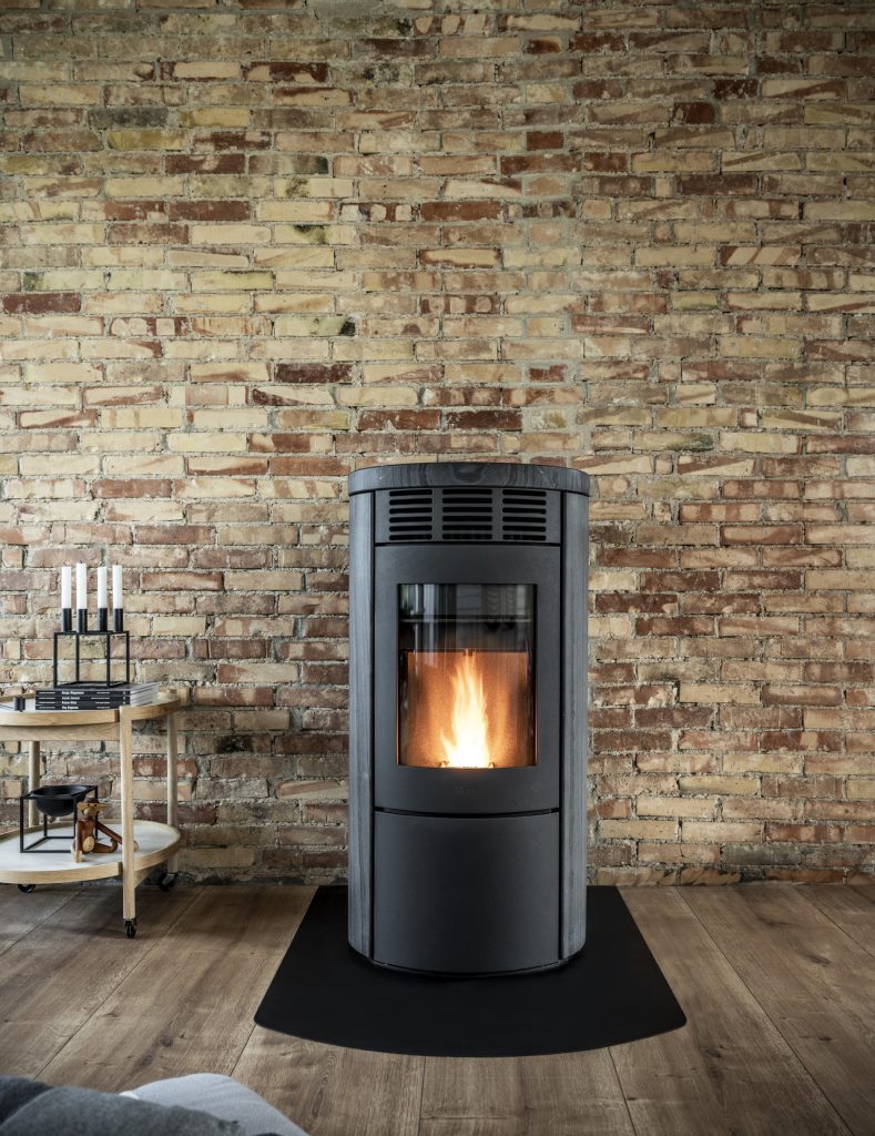 En modern svart Heta vedspis med synlig eld brinner framför en rustik tegelvägg på ett trägolv, med ett litet bord med levande ljus till vänster.