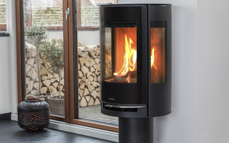En modern Aduro svart braskamin med glasfront och sidopaneler visar en ljus eld inuti. Staplade vedträn syns på utsidan genom stora glasdörrar, bredvid en lykta på golvet.