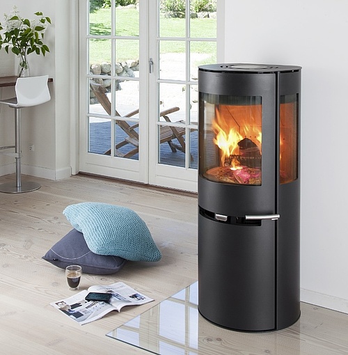 Aduro Kaminer: En komplett guide till danska designkaminer 3 I det ljusa vardagsrummet står en modern svart braskamin från Aduro med synlig eld. I närheten finns två kuddar, ett glas kaffe och en tidning på golvet. Stora fönster ger utsikt över uteplatsen och trädgården utanför.