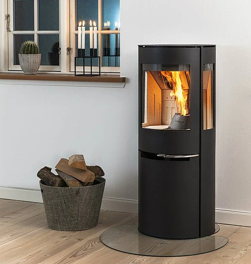 Aduro Kaminer: En komplett guide till danska designkaminer 4 En modern Aduro svart braskamin med flammor inuti står på en rund glasbas i ett mysigt rum. Veden förvaras i en grå hink i närheten, med levande ljus och en växt på fönsterbrädan i bakgrunden.