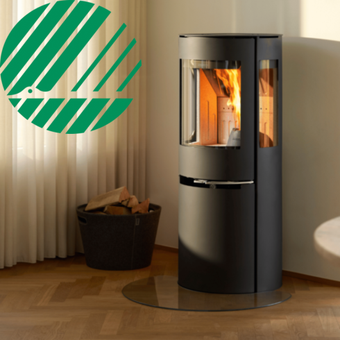 Aduro Kaminer: En komplett guide till danska designkaminer 5 En modern cylindrisk Aduro vedspis med synlig eld brinner intill ett fönster med vita gardiner. En korg med ved står bredvid och Svanens logotyp syns i det övre vänstra hörnet.