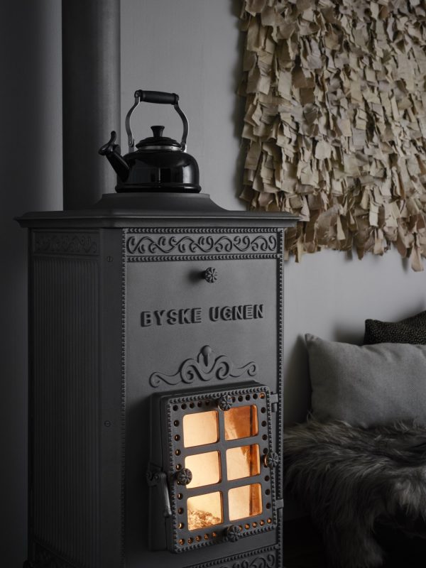 En svart vedspis av gjutjärn från Byskeugnen med BYSKE UGNEN på framsidan, en svart vattenkokare på toppen och en glödande eld inuti. En mysig interiör med en texturerad väggbonad och en pälsbeklädd bänk sitter bredvid den.