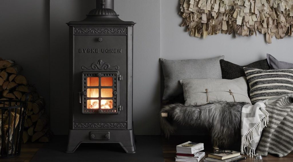 En svart vedspis i gjutjärn från Byskeugnen med en brasa som brinner inuti står bredvid en mysig sittgrupp med grå kuddar, en pälspläd, böcker och staplad ved mot en ljusgrå vägg som pryds av hängande tygkonst.
