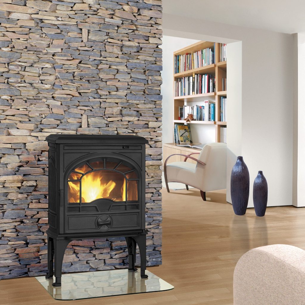 Dovre Kaminer: Norsk Gjutjärnstradition möter Modern Design 4 Ett mysigt vardagsrum har en svart vedeldad spis från Dovre mot en stenvägg med en eld som brinner inuti. Rummet har trägolv, vita väggar, bokhyllor, två blå vaser och moderna vita stolar.