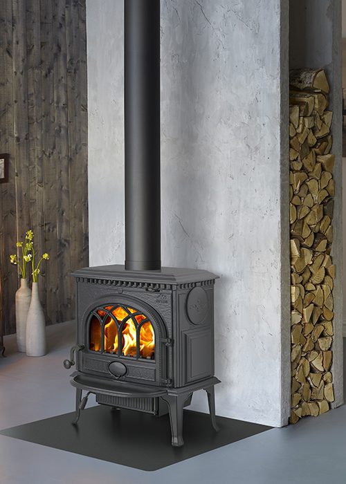Varför välja en Jötul? Mer än bara värme 4 En svart vedspis av gjutjärn från Jötul med ett högt skorstensrör står på en svart matta framför en vit vägg, med ved prydligt staplad i en nisch bredvid och en vas med gula blommor i bakgrunden.