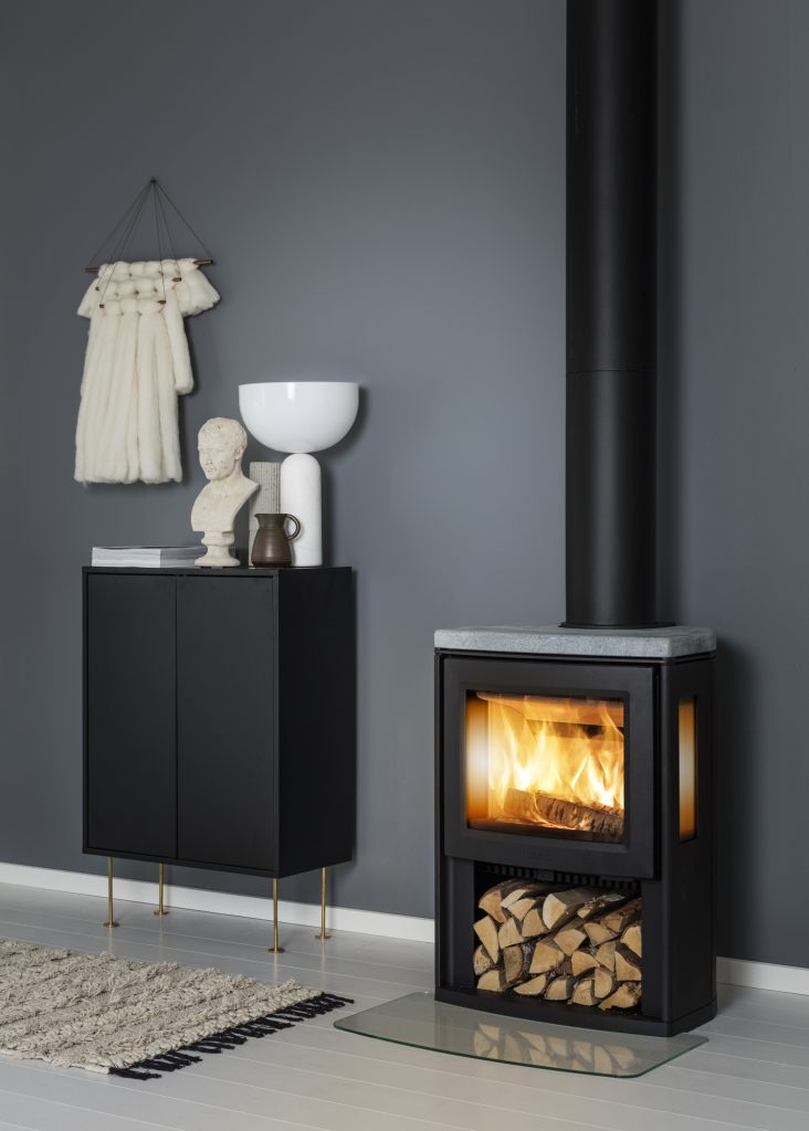 Ett modernt vardagsrum med en snygg vedeldad kedjekamin, en stapel ved nedanför och ett skåp med inredningsdetaljer. Den mörkgrå väggen kontrasterar snyggt mot det ljusa trägolvet och den lilla mattan.