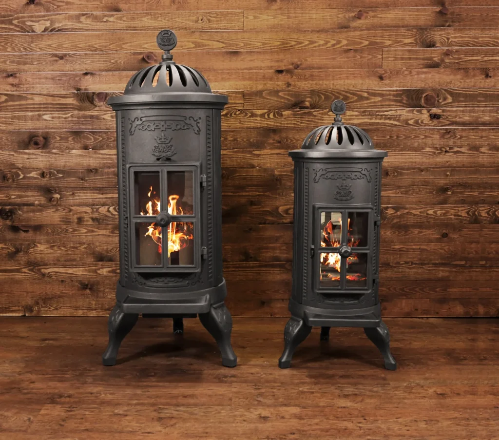 Två vedspisar av svart gjutjärn i vintagestil från Westbo med glasdörrar och synliga lågor inuti står på ett trägolv mot en vägg med träpanel. Den ena kaminen är större än den andra.