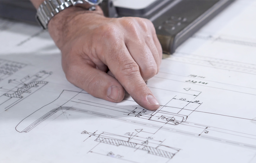 Varför välja en Jötul? Mer än bara värme 2 Närbild av en hand som pekar på tekniska ritningar och mätningar på en ritning för ett Jötul-projekt, som visar detaljerade tekniska eller arkitektoniska planer. Personen bär en klocka.