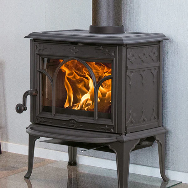 Varför välja en Jötul? Mer än bara värme 5 En svart vedspis i gjutjärn från Jötul med glasdörr avslöjar glödande lågor inuti och står på en reflekterande yta mot en ljus vägg.