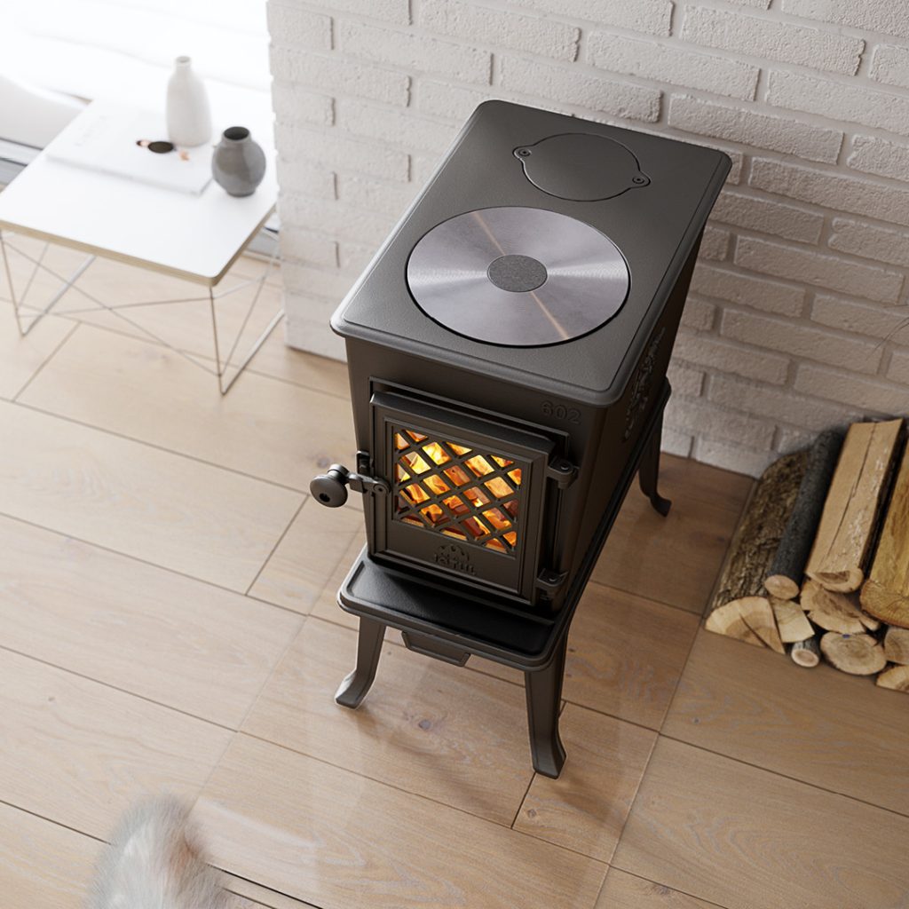 Varför välja en Jötul? Mer än bara värme 3 En svart Jötul-vedspis med en glasdörr som visar en eld inuti står på ett ljust trägolv bredvid staplade vedträn och en vit tegelvägg. I närheten står ett vitt sidobord med keramik.