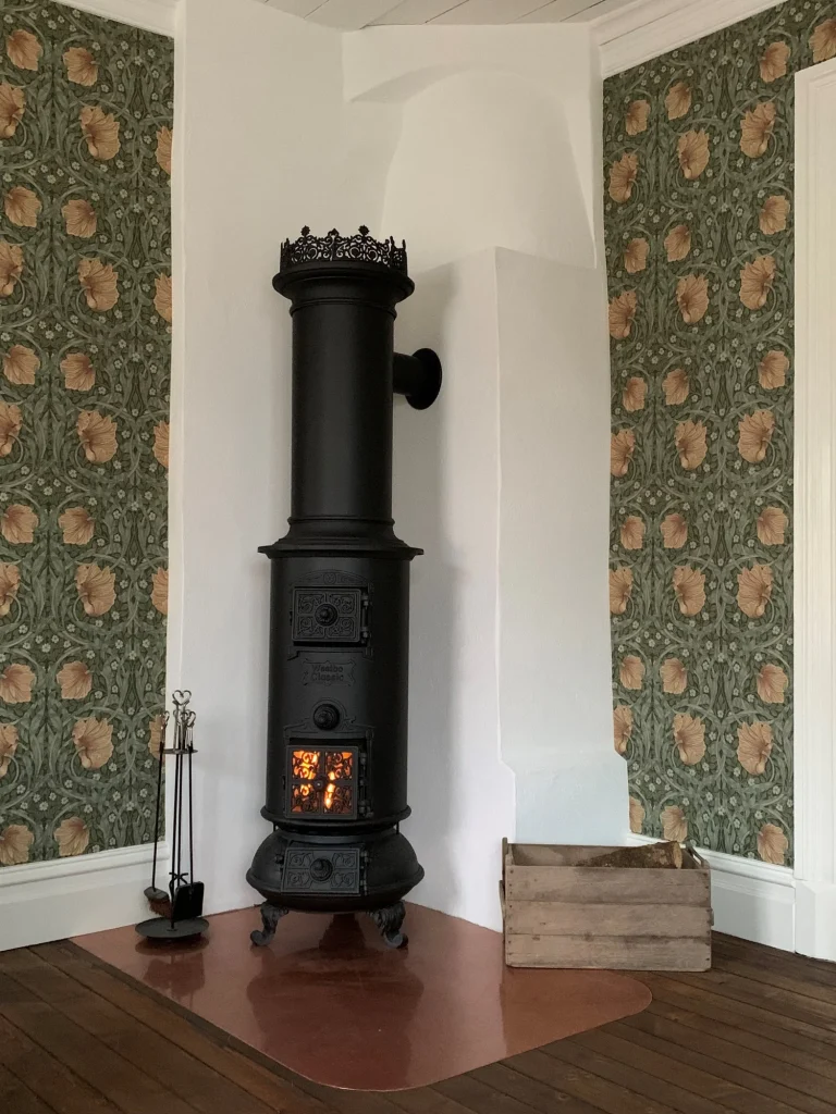 En vintage svart Westbo gjutjärnskamin står i hörnet av ett rum med blommig tapet. Kaminen är tänd och elden syns inuti. Bredvid den finns en uppsättning eldstadsverktyg och en tom trälåda.