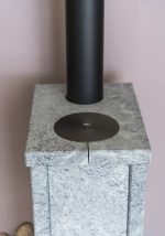 Närbild på vedspisen Norsk Kleber Otta 102 i grå sten, med skorstensrör i svart metall och en rund metallplatta på toppen, mot en ljusrosa vägg.