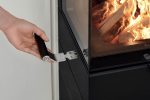 En hand använder ett metallverktyg med svart handtag för att öppna glasdörren till den moderna vedeldade kaminen Leda Corna PS, och avslöjar en brinnande eld inuti.