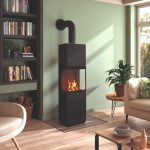 I ett modernt vardagsrum står den svarta braskaminen Leda Corna PS mot en grön vägg, med eld inuti. I rummet finns en bokhylla, en fåtölj, en krukväxt, ett fönster och ett soffbord på trägolv.