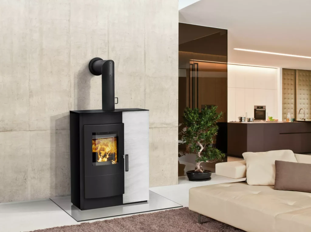 En modern Rika braskamin med synlig eld är installerad mot en betongvägg i ett stilrent, minimalistiskt vardagsrum med beige möbler, en krukväxt och ett öppet kök i bakgrunden.