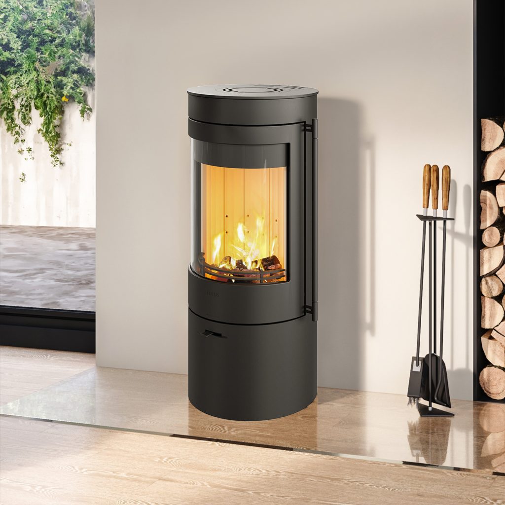 Lotus Kaminer: En guide till funktion & design 2 En modern svart Lotus-vedspis med glasfront som visar en ljus eld inuti, placerad i ett minimalistiskt rum med vedträn staplade i närheten och eldstadsverktyg stående till höger.