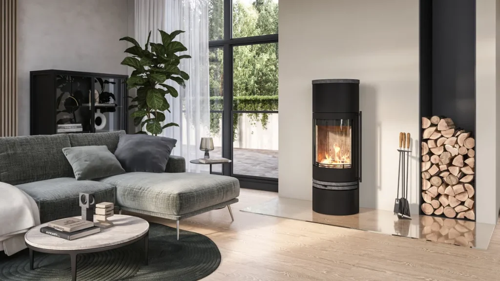 Modernt vardagsrum med grå soffa, grön växt, Lotusinspirerat runt soffbord och en tänd svart vedspis bredvid veden vid stora fönster med utsikt över utomhusmiljön.