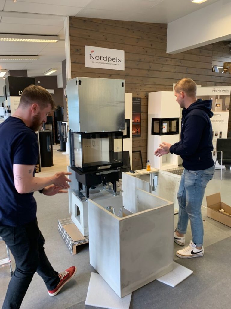 Två män monterar en modern Nordpeis-eldstad i ett utställningsrum med träväggar och montrar. Den ena mannen håller i ett verktyg, medan den andra står bredvid. På en skylt ovanför står det Nordpeis.