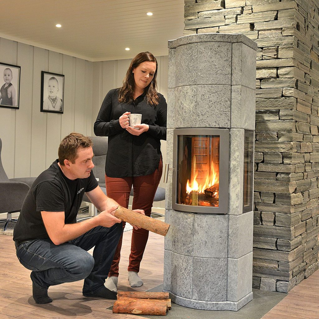 En man står på knä och lägger ved i en Norsk Kleber-stenkamin medan en kvinna står bredvid och håller i en mugg, båda i ett mysigt vardagsrum med familjefoton på väggen.