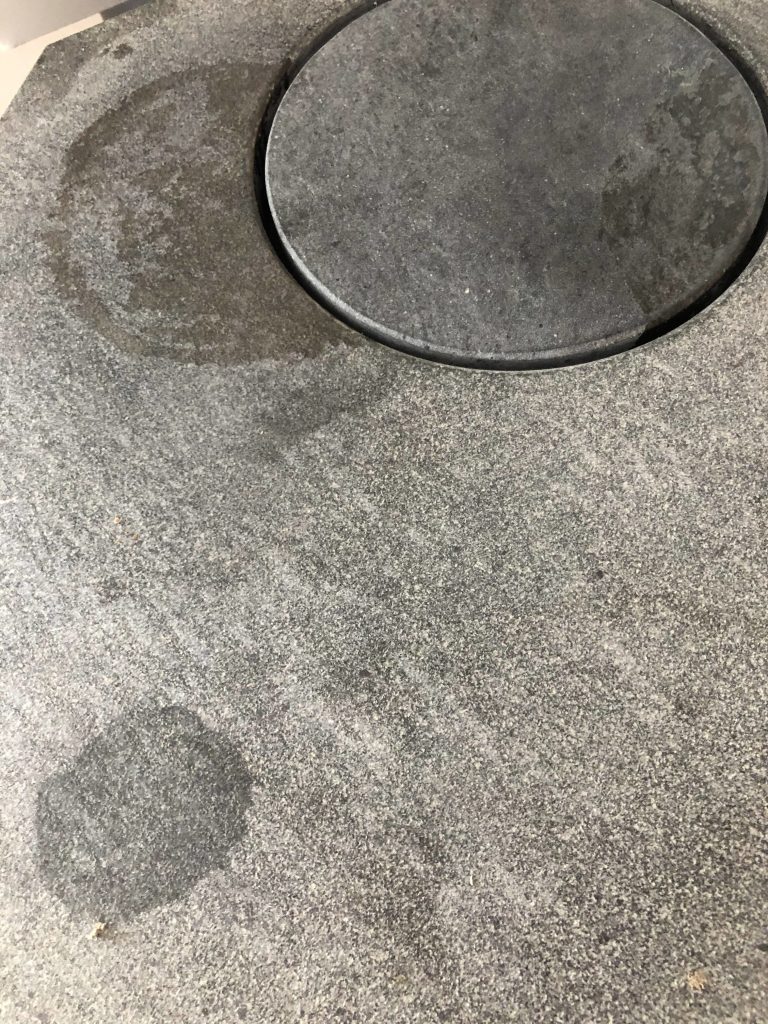 En närbild av en grå stenyta med en cirkelformad upphöjd del och flera oregelbundna mörkare våta fläckar utspridda över den, vilket visar detaljer att tänka på när du behöver rengöra täljsten.