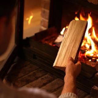 En person håller i en vedkubbe och förbereder sig för att lägga den i en tänd eldstad med synliga lågor och glödande brasa.