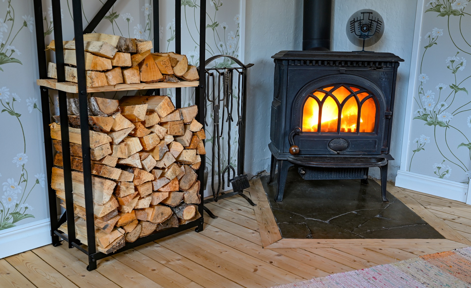 En svart vedeldad kamin, utformad efter en braskamininstallation, står på en kaklad eldstad bredvid en metallhylla med ved. Rummet har trägolv, blommig tapet och synliga eldstadsverktyg.