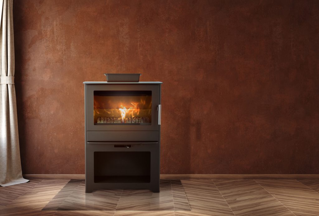 En modern vedeldad spis med kamin kokplatta, glasdörr och en liten svart kruka ovanpå, mot en brun strukturerad vägg och ljust chevron trägolv, med beige gardiner delvis synliga till vänster.