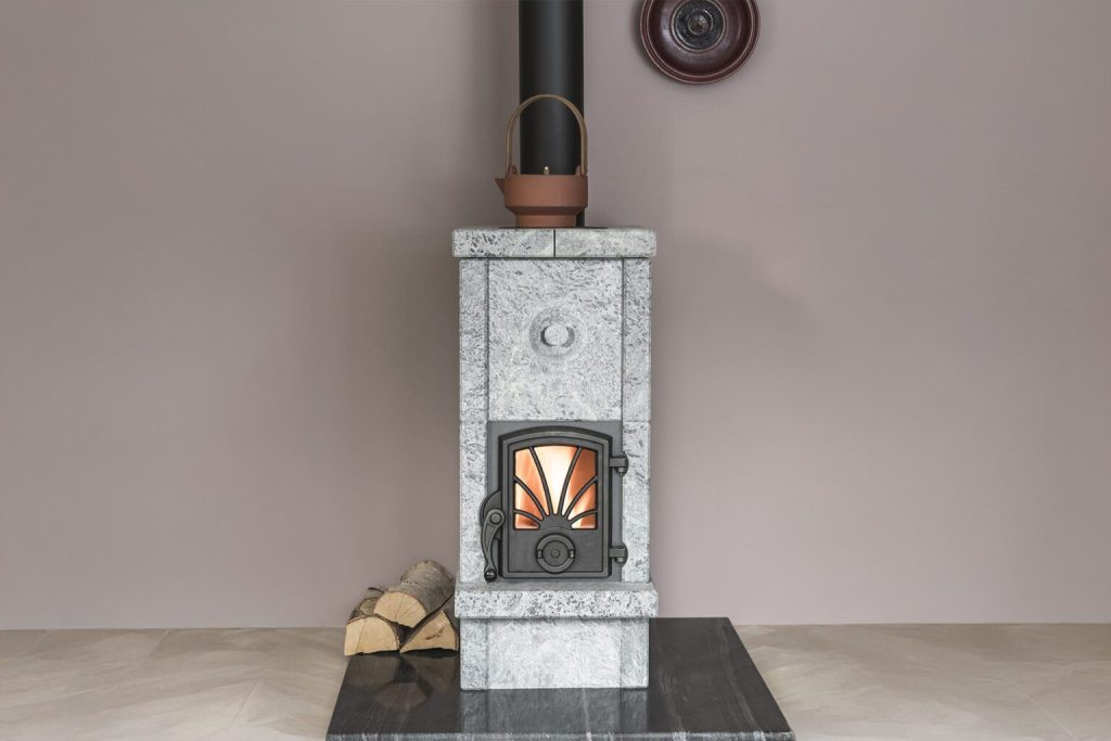 En modern vedeldad stenkamin med en tänd brasa inuti, ett svart skorstensrör, en brun vattenkokare på sin kamin kokplatta och en liten stapel ved bredvid, placerad mot en vanlig ljus vägg.