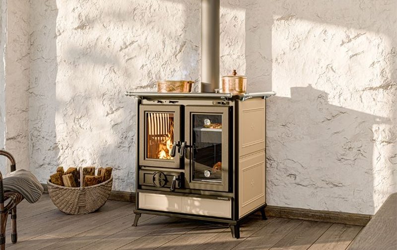 En vedeldad kamin i krämfärgat och svart, betraktad av många som bästa vedspisen, står mot en vit texturerad vägg; två kopparkrukor står ovanpå och en korg med ved vilar i närheten.