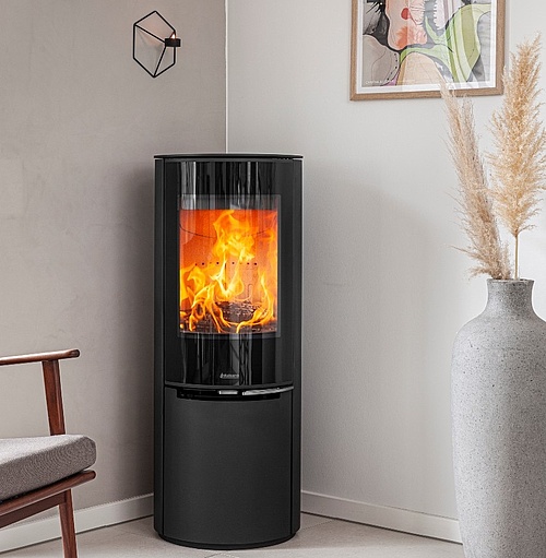 En modern rund kamin svart vedeldad spis med en glasdörr som visar ljusa lågor är placerad i ett mysigt hörn nära en beige stol, vit vas med pampasgräs och väggkonst på en ljus vägg.