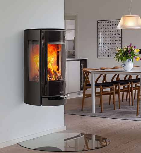 En modern svart rund kamin väggmonterad vedspis med en eld som brinner inuti, placerad i ett minimalistiskt vardagsrum med ljusa trägolv och en eldstad i glas. I bakgrunden skymtar en matplats med träbord och stolar.