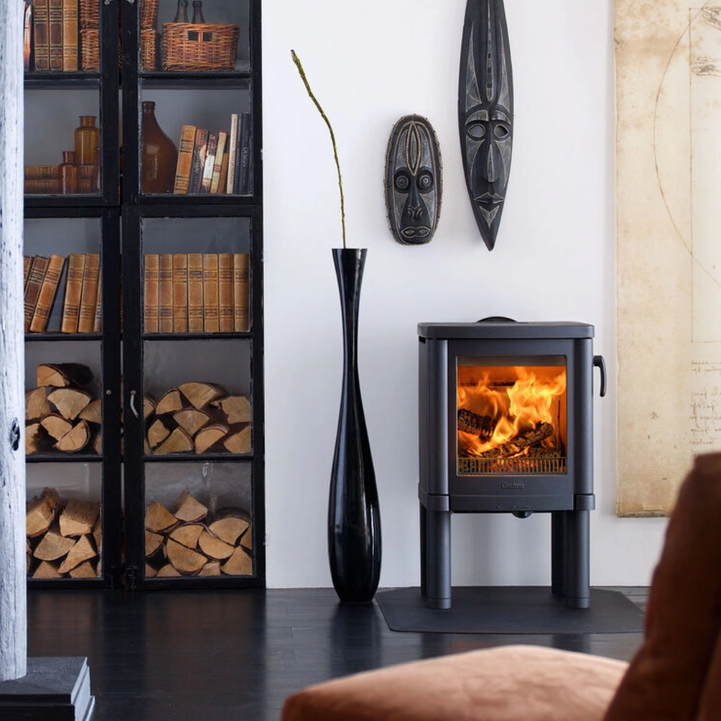 Ett mysigt vardagsrum med en liten kamin, staplad ved, en svart vas med en enda gren, bokhyllor och två afrikanska masker som hänger på väggen. Varma lågor syns inuti kaminen.