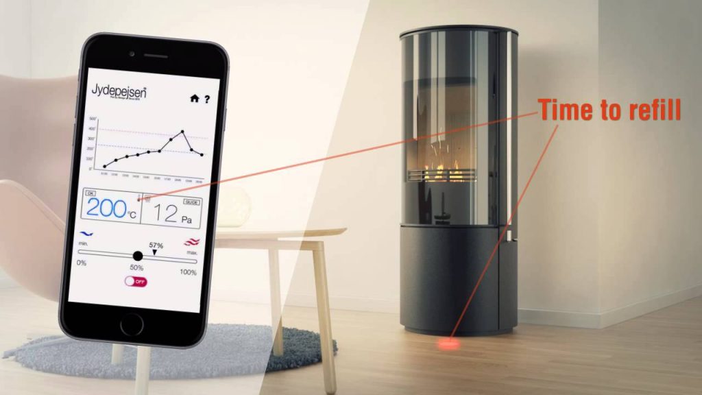 En modern vedspis från Jydepejsen i ett vardagsrum är ansluten till en smartphone-app som visar dess status. En varning på bilden säger Dags att fylla på, med en linje som pekar mot kaminens bränsleutrymme.