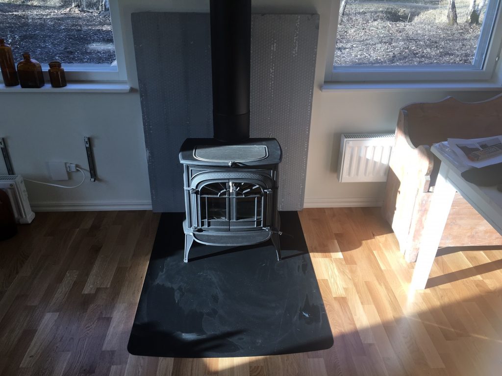 En svart vedspis står på en svart matta över ett ljust trägolv, med en värmesköld av metall bakom sig för avstånd till brännbart material. Solljuset skiner in genom fönstren i närheten, medan bruna flaskor och ett litet bord syns i rummet.