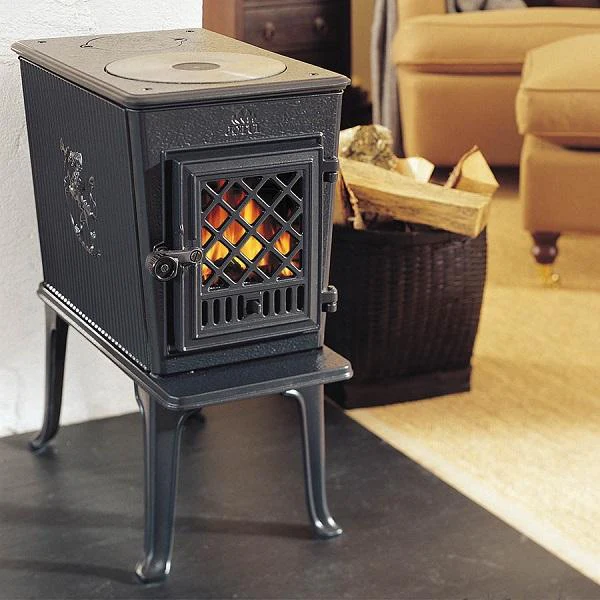 En svart vedeldad gjutjärnskamin med gallerlucka och kamin kokplatta står på en eldstad, med en korg med ved och beige möbler som syns i den mysiga vardagsrumsbakgrunden.