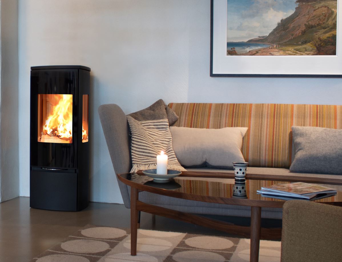 Ett mysigt vardagsrum med en randig soffa, kuddar, ett tänt ljus och en kopp på ett soffbord i trä, en mönstrad matta och en modern vedeldad kamin från Jydepejsen med lågor under en landskapsmålning på väggen.