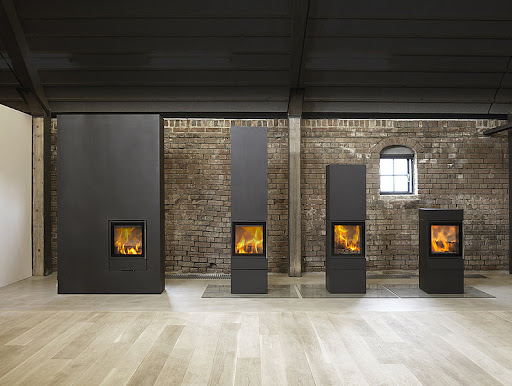 Fyra moderna svarta Jydepejsen-vedspisar i olika höjder står uppställda mot en tegelvägg med ett enda litet fönster, i ett rum med trägolv och mörkt tak.