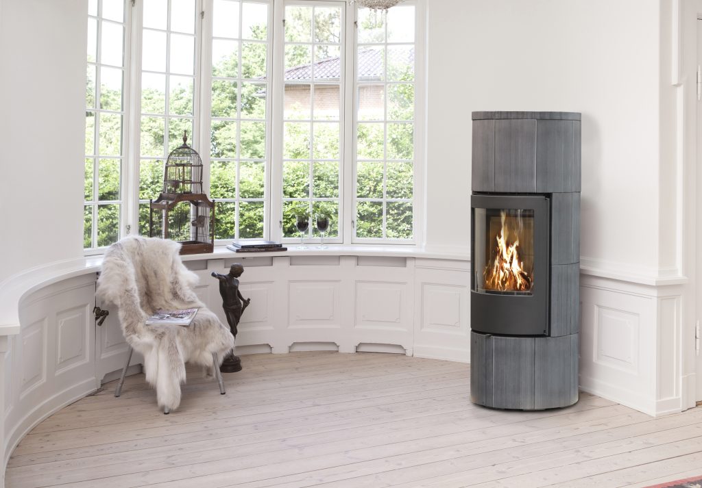 Ett modernt, minimalistiskt rum med stora fönster, en vedeldad kamin från Jydepejsen med synlig eld, en pälsbeklädd stol, ett litet bord, prydnadsföremål och ljusa trägolv. Naturligt ljus fyller det mysiga, inbjudande utrymmet.