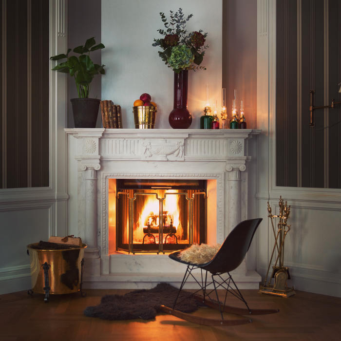 Ett mysigt vardagsrum med en tänd murspis i en vit utsmyckad spiselkrans, dekorerad med växter, böcker och levande ljus. En svart fåtölj med päls ligger på en matta i närheten, och till vänster och höger finns ved och eldstadsverktyg.