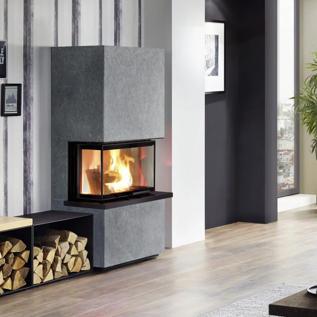 Ett modernt vardagsrum med en elegant, grå murspis som brinner starkt. Staplade vedträn vilar på en hylla i närheten och stora fönster släpper in naturligt ljus som lyser upp trägolvet.