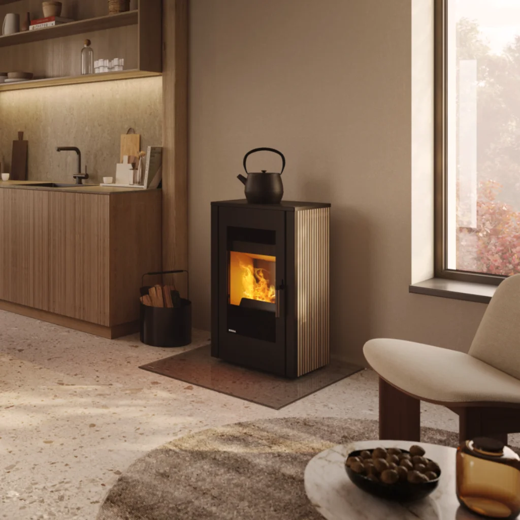 Ett mysigt modernt kök har en kamin kokplatta med en svart vattenkokare ovanpå, bredvid ett fönster. En ljus fåtölj och en skål med nötter står i förgrunden på ett stengolv.