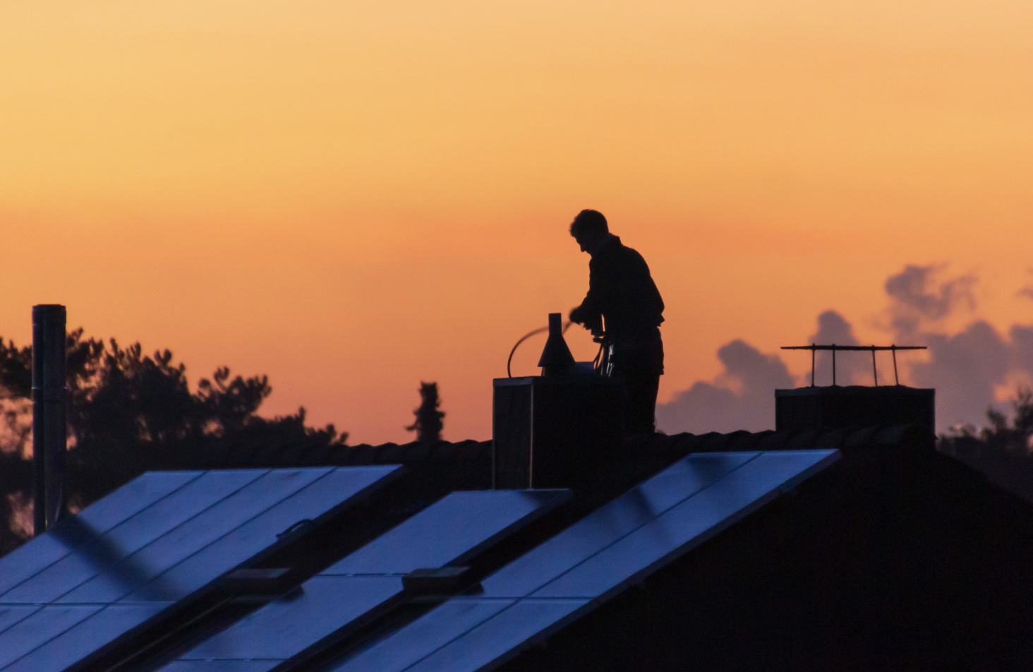 En silhuett av en person som arbetar på ett tak i solnedgången, kanske utför en brandskyddskontroll, med en orange himmel och trädtoppar i bakgrunden. Hustaken reflekterar det bleknande solljuset.
