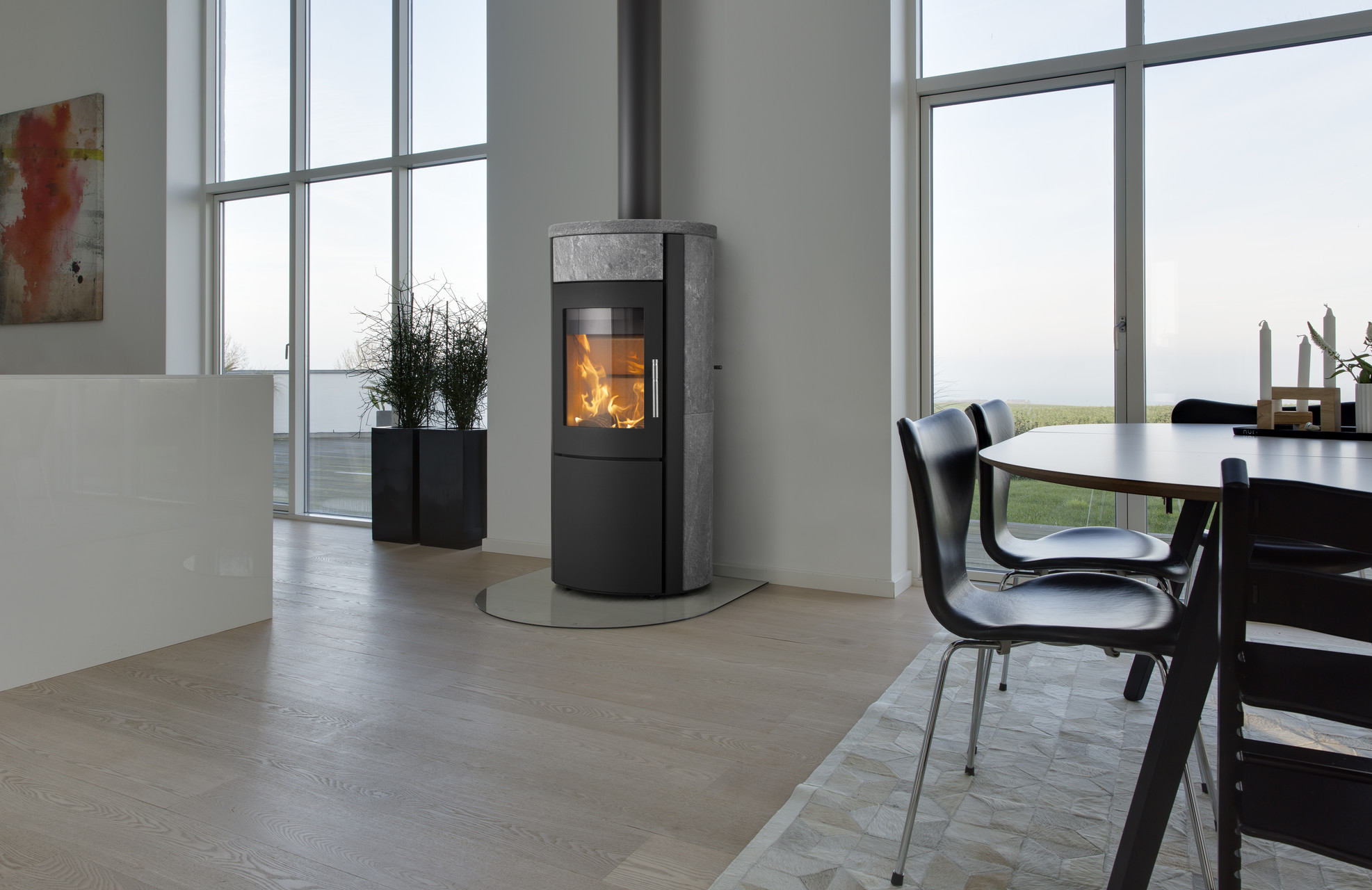 Ett modernt vardagsrum med stora fönster, ljusa trägolv, en svart vattenmantlad kamin med en synlig eld, ett runt matbord med svarta stolar och krukväxter vid fönstret.