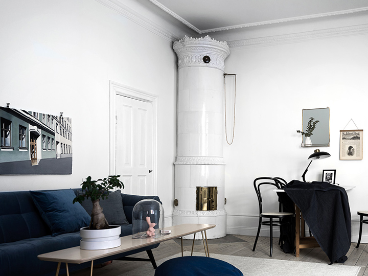 Ett stilrent vardagsrum med vita väggar, en hög vit kakelugn i hörnet, marinblå soffa, soffbord och en liten matplats med svarta stolar och ett bord draperat i en mörk duk. Väggarna pryds av konstverk.