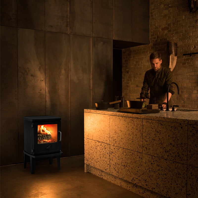 Morsø Kaminer: En guide till de danska gjutjärnsikonerna 2 En man lagar mat i ett svagt upplyst kök med bänkskivor i sten och tegelväggar. En liten svart vedeldad kamin med en eld inuti ger värme och ljus till det mysiga, moderna utrymmet.